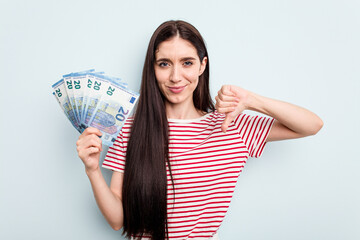 Young caucasian woman holding banknotes isolated on blue background showing a dislike gesture, thumbs down. Disagreement concept.