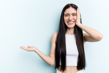 Young caucasian woman isolated on blue background holds copy space on a palm, keep hand over cheek. Amazed and delighted.