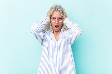 Middle age caucasian woman isolated on blue background covering ears with hands trying not to hear too loud sound.