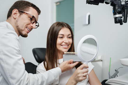 Young man dentist in white coat show veneers enamel color palette for beautiful patient woman
