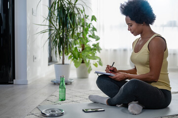 Young African-American woman writing in her notebook. Sitting at home by the window on a floor...