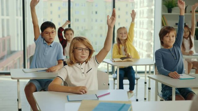 Elementary classroom of diverse bright children listening to the teacher giving a lesson. Everyone raises hand knowing the right answer. Brilliant kids in school learning science, creative thinking.