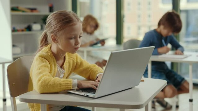 Side view of the girl student using laptop for studying purposes at school in the class and other pupils on the background. People and technology concept.