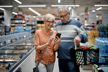 Senior couple using smart phone while buying groceries in supermarket.