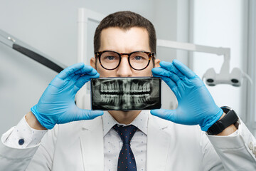 Male dentist cover his mouth with a smartphone with X-ray of his teeth on a screen of device.