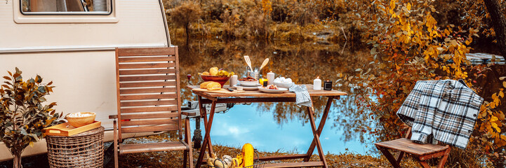 trailer of mobile home or recreational vehicle stands on shore of pond in camping in autumn near table set, concept of family local travel in native country on caravan or camper van and camping life - obrazy, fototapety, plakaty