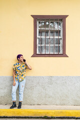 Obraz na płótnie Canvas vertical portrait of a bisexual woman leaning against a yellow wall with a window talking on a cell phone dressed in male clothing and smiling