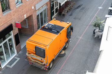 Fototapeta premium Top above view of small sweeping vacuum cleaner machine equipment removing dust rubbish city street sidewalk paved road near apartment condo building. Special municipal sweeper vehicle cleaning town