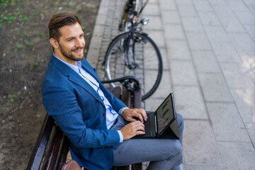 Obraz premium Portrait of happy entrepreneur working on laptop\tablet while sitting on park bench. Bicycle in blurred background.