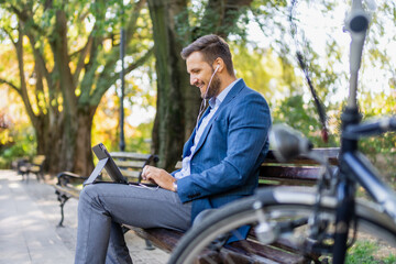 Fototapeta premium Caucasian male entrepreneur freelancer sitting outdoors in city park a bench talking online a video call using laptop or tablet. Business man office worker working on urban street background.