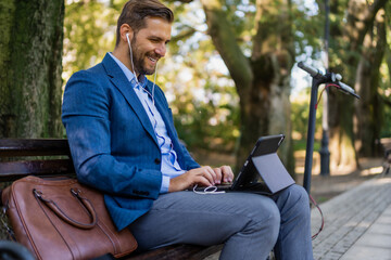 Caucasian male entrepreneur freelancer sitting outdoors in city park a bench talking online a video call using laptop or tablet. Business man office worker working on urban street background.