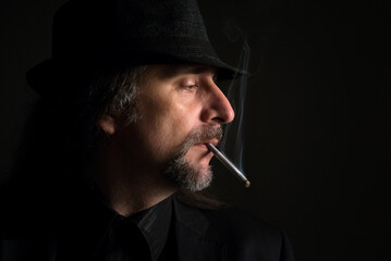 Portrait of middle aged man wearing black clothes and black hat smoking cigarette in the studio
