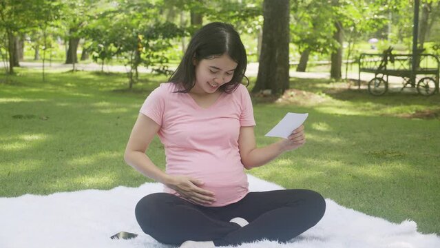 Happiness young asian woman pregnant sitting touch belly and looking photo with positive in the park, mother and health of pregnancy, happy of parenthood, one person, lifestyles and family concept.