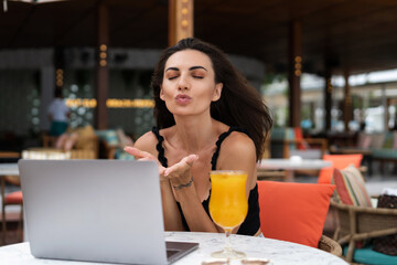 Beautiful young female freelancer working with a laptop in a street cafe, making video call, sending kiss, calling friends, family