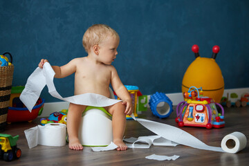 Cute smiling baby boy sitting on chamber pot with toilet paper rolls. Potty training. Domestic life
