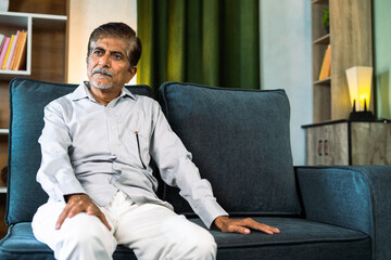 Thoughtful grandfather sitting on sofa at home showing with copy space - concepts of financial problems, loneliness and mental illness