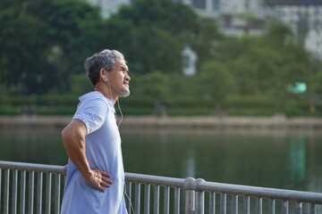 asian senior man with grey hair and beard wearing headphones for listening music is warming up in the city park before workout, concept elderly people lifestyle, healthcare, happiness, well being