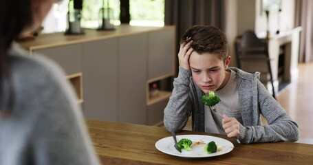 Frustrated boy told to eat healthy vegetables that he hates eating by his mom at meal time on his dinner plate. Mother stands in the kitchen to watch her unhappy and upset child finish his broccoli.