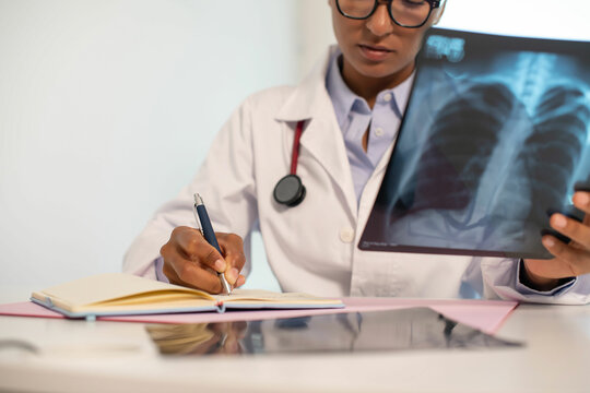 Close-up of pulmonologist taking notes in office. African American doctor wearing glasses holding x-ray picture of human lungs. Pulmonology concept