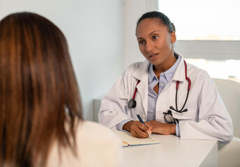 Fototapeta premium Confident young doctor writing down anamnesis of female patient. African American general practitioner sitting at table and talking to woman in her office. Visit to doctor concept