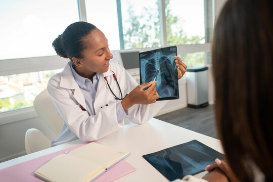 Pulmonologist showing x-ray picture and explaining diagnosis to female patient. Young African American doctor sitting at table and consulting woman. Medical consultation, pulmonology concept