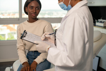 Smiling female patient asking questions of doctor in clinic. Female general practitioner talking to young African American woman and filling in questionnaire. Medical exam concept - obrazy, fototapety, plakaty
