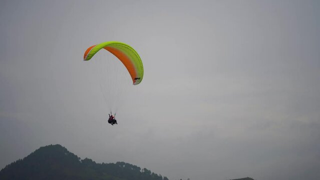 zoomed shot showing para gliding people with a bright orange yellow glider in the middle of hills mountains near nanital bir billing showing a popular adventure sport for tourists