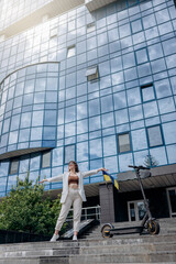 Beautiful young woman in sunglasses and white suit standing with her electric scooter near modern building and looking away