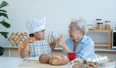 Asian family grandmother and grandchild have fun baking cookies at home kitchen together, little cute granddaughter with chef hat and apron is thumbs up after eating homemade cookie from Grandma