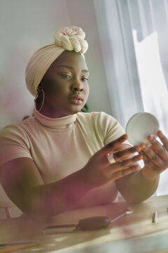 Young African American Woman In Traditional Beige Turban Examining Makeup In Front Of Mirror