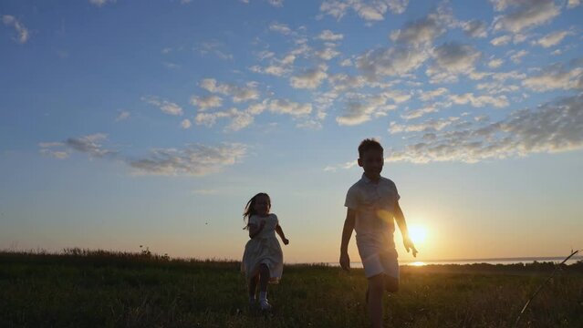 Happy children enjoy playing together against blue sky. Kids silhouettes run on evening meadow at back sunset. Siblings enjoy walking on nature