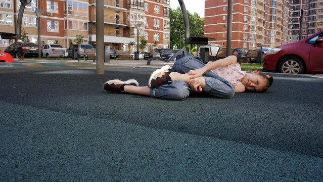 A sad girl is lying on the rubber covering of the playground holding her bruised leg.