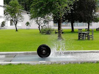Dynamic sculpture featuring a spinning car wheel suspended above water, spraying and splashing in a...