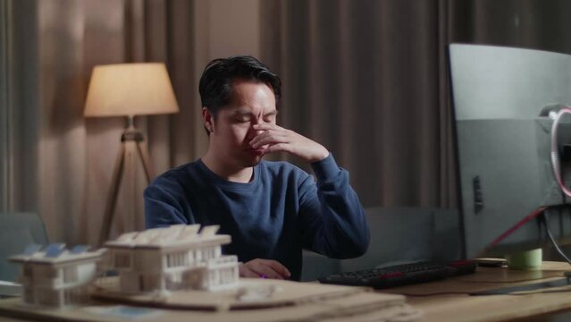 Asian Male Engineer With The House Model Having A Headache While Working On A Desktop At Home
