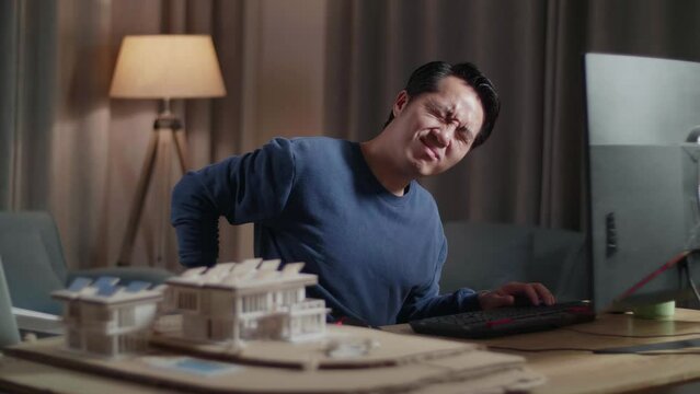 Asian Male Engineer With The House Model Having A Headache While Working On A Desktop At Home
