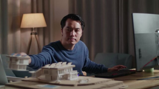 Asian Male Engineer Holding And Looking At The Small House Model With Solar Panel Before Comparing It To The Pictures On A Desktop At Home
