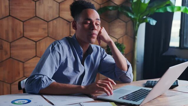 Young professional African American man fills out tax forms using laptop and waits for confirmation of correctness entered data looks at monitor sits in office at table with papers and calculator
