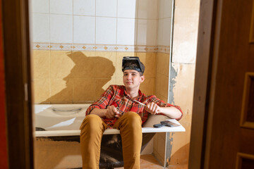 a young man, a plasterer or painter, sits in a bathtub with a spatula in his hands and looks at the...