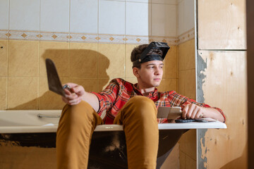 a young man, a plasterer or painter, sits in a bathtub with a spatula in his hands and looks at the...