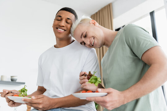 Low Angle View Of Multiethnic Gay Couple Smiling While Holding Fresh Salad In Kitchen