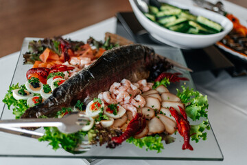  beautifully arranged platter featuring a whole baked fish with fresh vegetables and seafood, served on a glass plate.