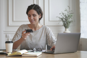 Young smiling italian woman in casual clothes looking at credit card and typing on laptop