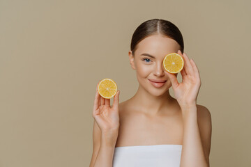 Lemon for beauty and health. Beautiful healthy young woman covers eye with half of lemon enjoys spa treatment wrapped in bath towel isolated over beige background copy space for your promotion