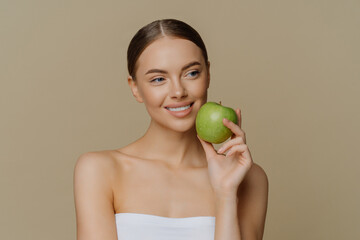 Thoughtful charming European woman holds apple near face smiles gently has white perfect teeth...