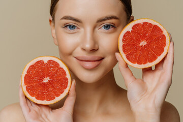 Close up shot of healthy lovely woman holds halves of grapefruit near face has healthy glowing skin...