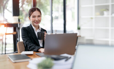 Smiling Asian businesswoman holding a coffee mug and laptop at the office. Looking at the camera.