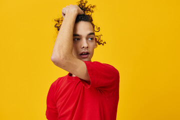 Joyful funny Caucasian young man in red t-shirt touches hair curls posing isolated on over yellow studio background. The best offer with free place for advertising. Emotions for everyday concept