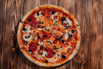 Tomato-based pizza with mozzarella cheese, hunting sausages, salami, olives, mushroom sauce. On a wooden background