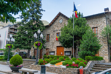 Sant Julia de Loria Town Hall in Andorra, a stone building with the Andorran flag on a mast and a garden with trees and flowers in summer, with the mountains behind and closed wooden doors