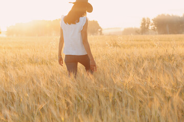 Woman farmer in cowboy hat walking with hands on ears at agricultural field on sunset.